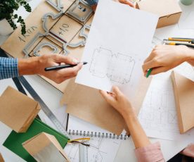 Top view of box designers desk. Man and woman hands at work. There is pencil pointing and blueprint scetch holding.