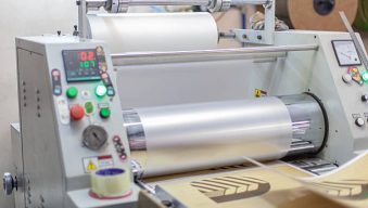 Close-up photo of commercial roll laminator in print shop applying protective film to printed cardboard sheets.